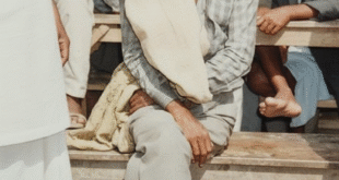 ডঃ বশিষ্ঠ নারায়ণ সিং একটা সাদা রঙের আসনে বসে আছেন মানসিক ভারসাম্য হারিয়ে। চারপাশে লোকের ভিড়। তাঁর লম্বা, সাদা দাড়ি ও চুল, চিন্তিত মুখে তাকিয়ে ক্যামেরার দিকে।